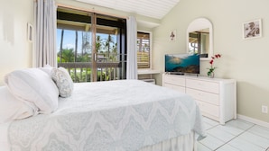 A bedroom with a bed, a TV, a dresser, and a balcony with a view of palm trees.