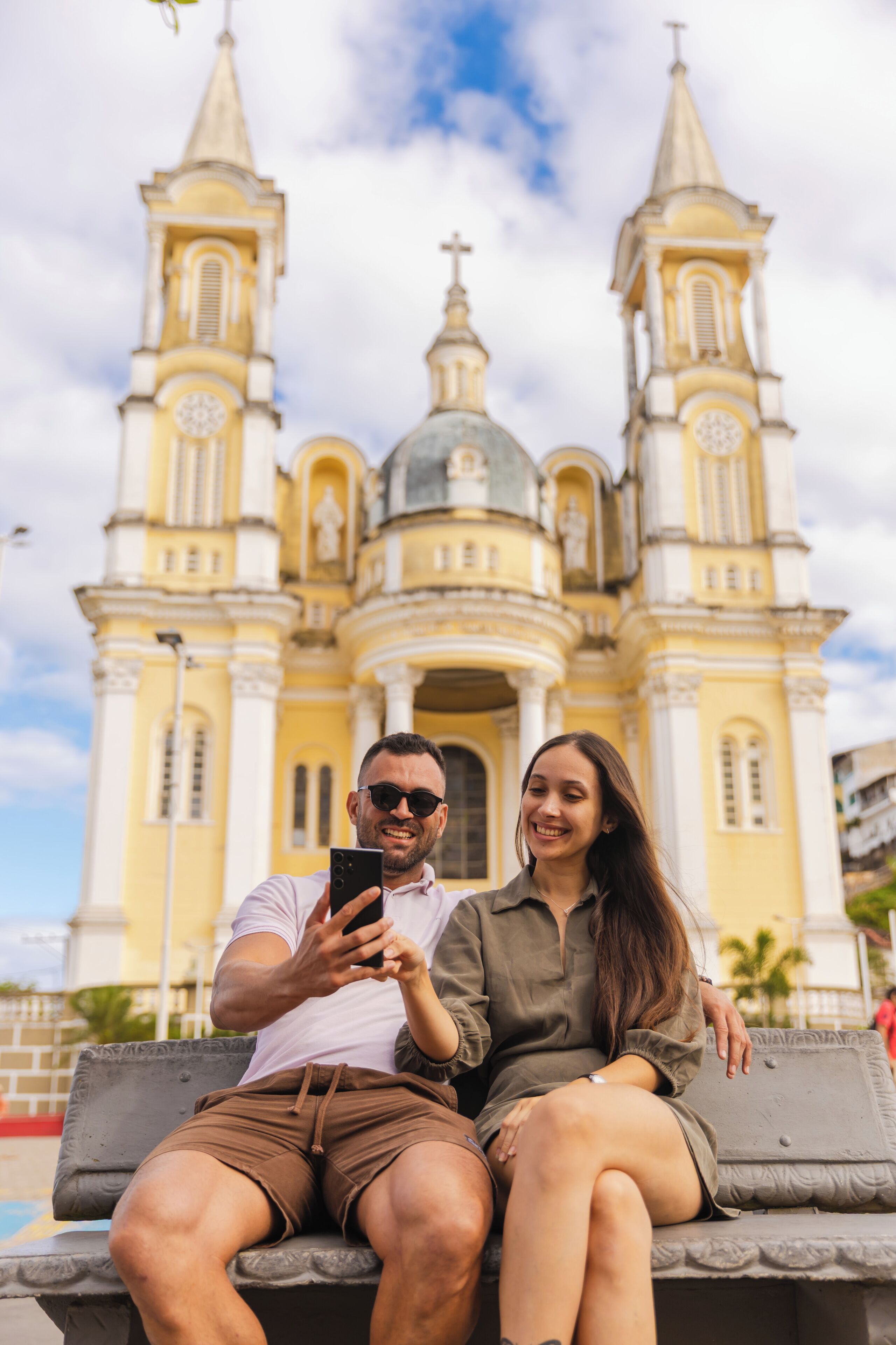 Una pareja sentada en un banco frente a una iglesia amarilla grande con dos torres.