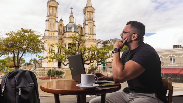 Una persona sentada a una mesa con una computadora portátil, una taza y una mochila. Al fondo, se aprecia una iglesia grande con varias torres.