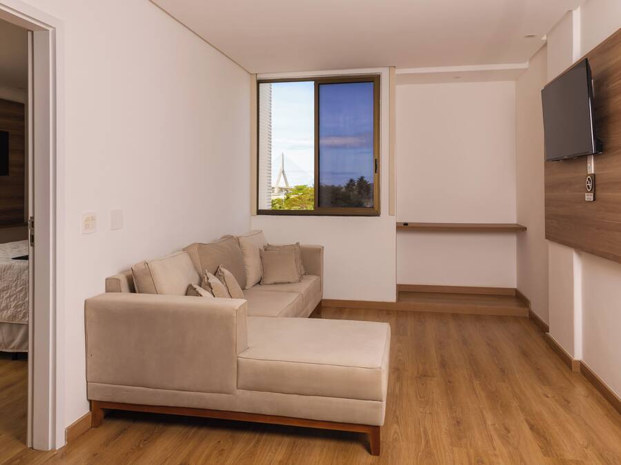Una sala de estar moderna con un sofá seccional color beige, un televisor de pantalla plana montado en una pared de madera y un ventanal con vista a un edificio y árboles.