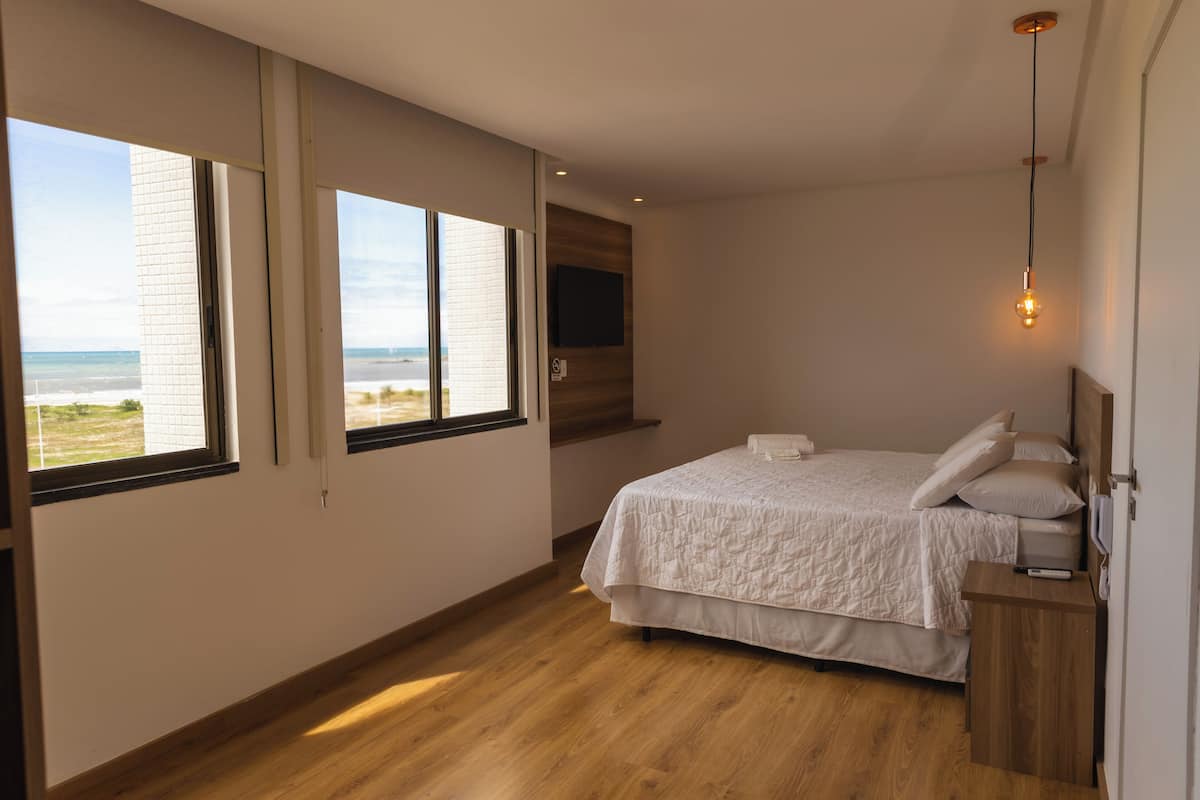 A bedroom with a large bed, a TV mounted on the wall, and a view of the beach through the windows.