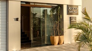 Exterior del Hotel Ilhéus Praia, con un diseño moderno, que presenta una entrada de vidrio, plantas en macetas y un pavimento empedrado.