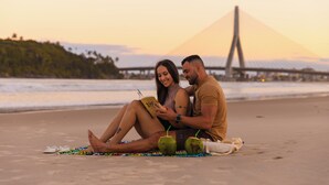 Una pareja sentada en la playa, con un puente al fondo.
