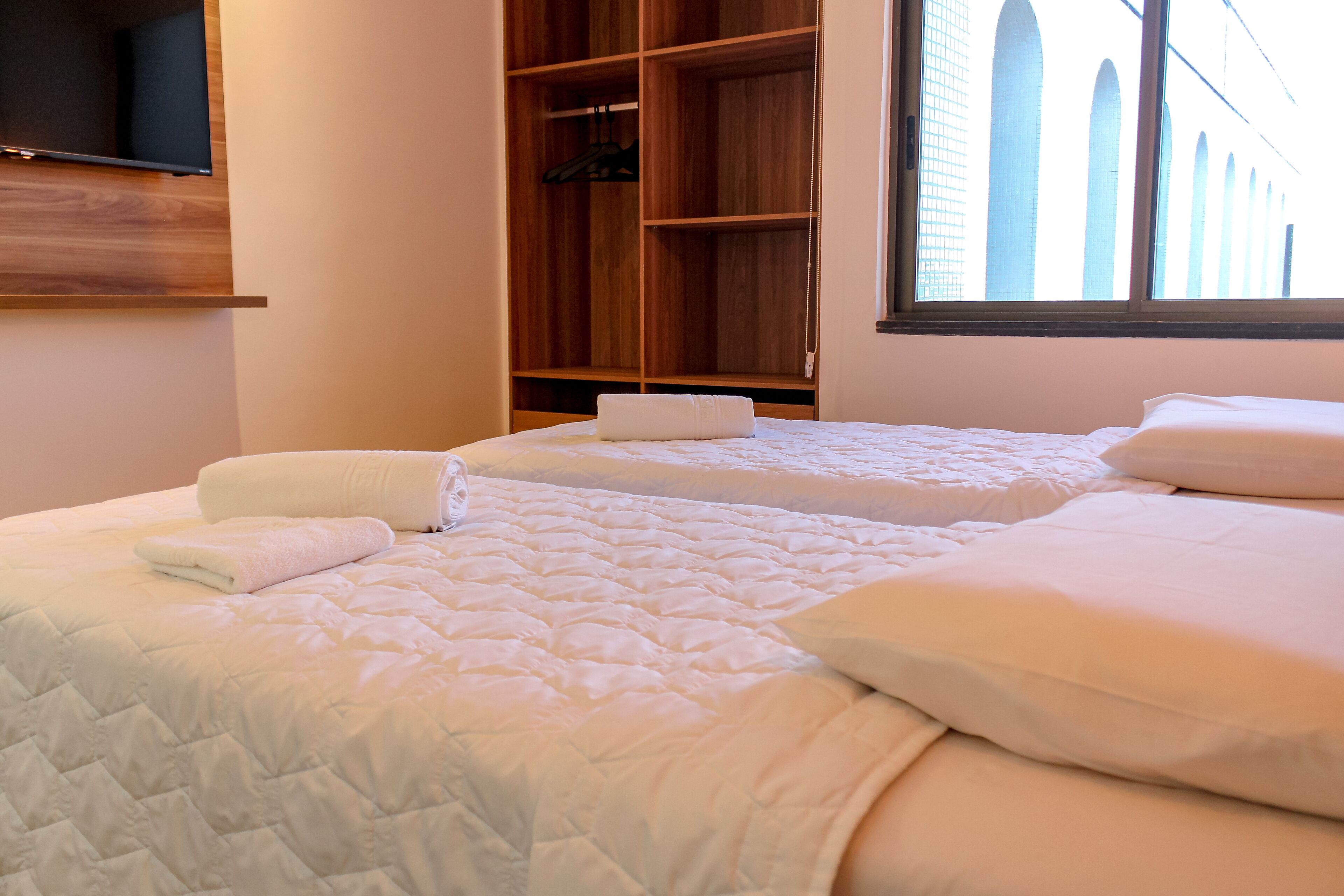 A hotel room with two beds, a wooden wardrobe, and a TV mounted on the wall.