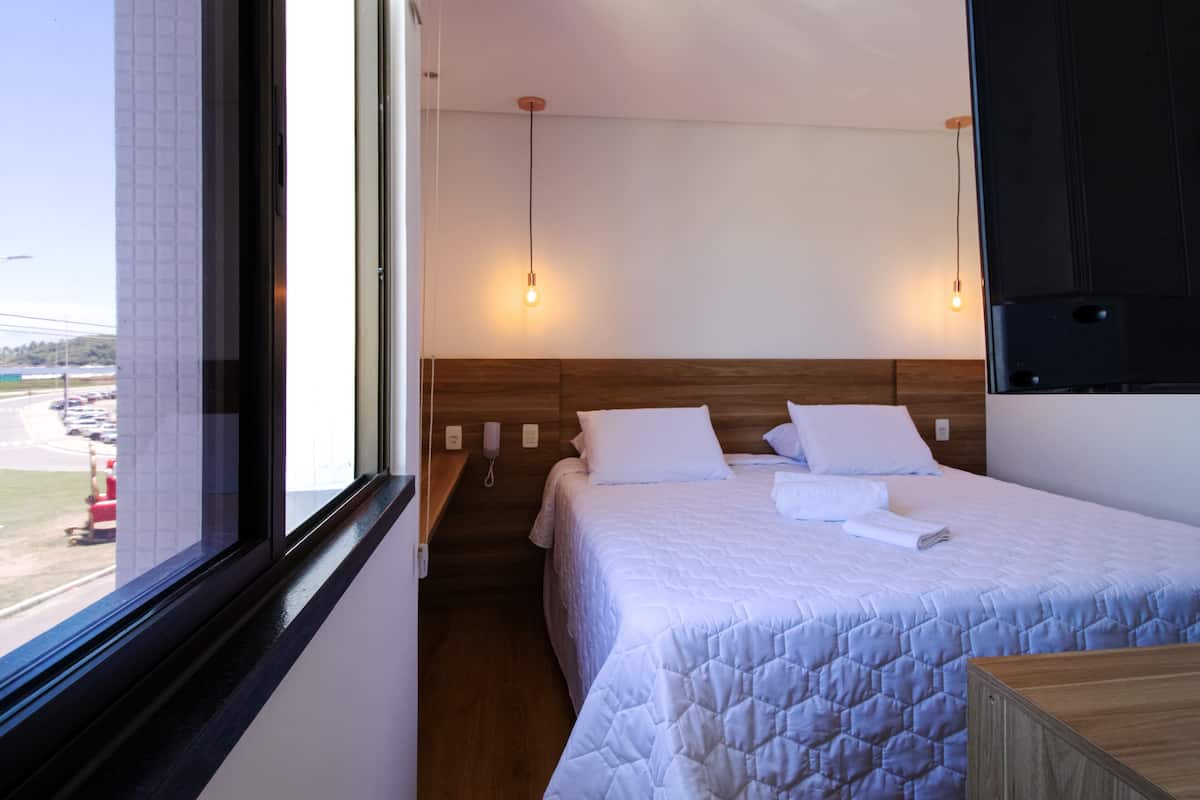 A hotel room with a large bed, a wooden headboard, a bedside table, a nightstand, a window with a view of the outside, and a wall-mounted TV.