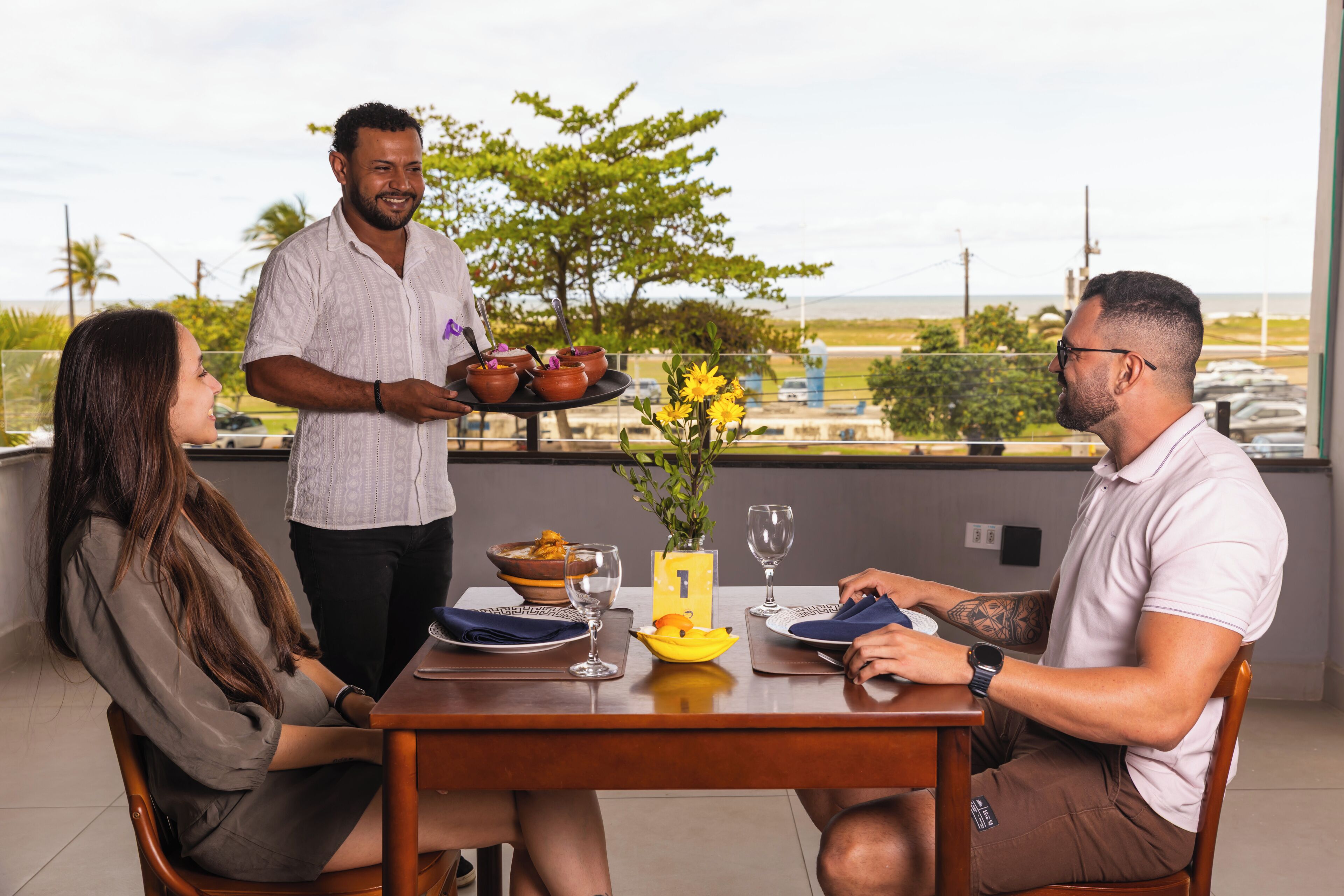 Una mesa de comedor con un camarero sirviendo comida, un florero con flores y vistas al océano.