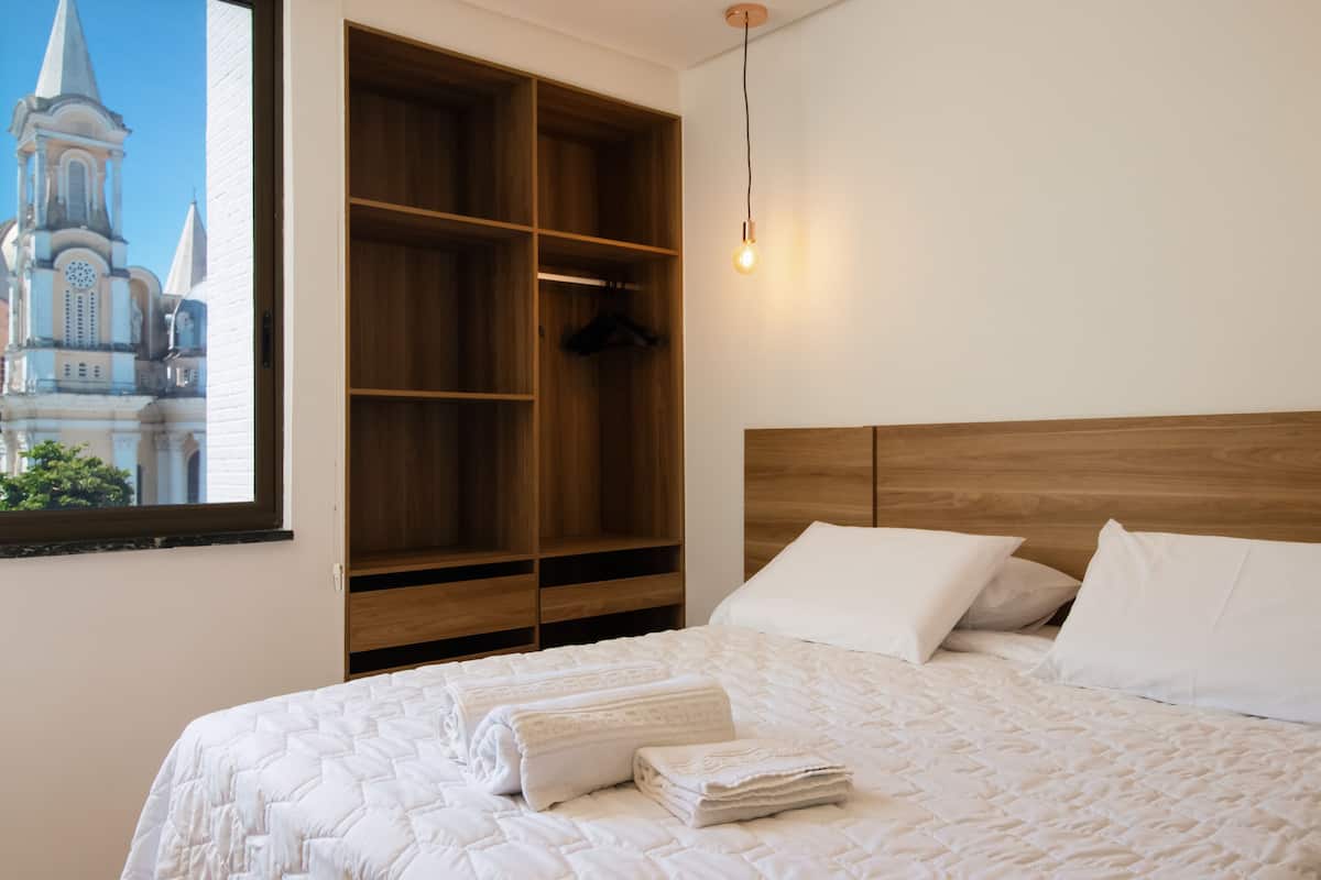 A neatly made bed with white linens and a view of a church through a window.