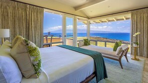 A hotel room with a large bed, two chairs, a view of the ocean, and a balcony.