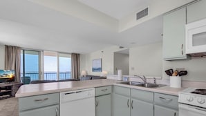 Een moderne keuken met witte apparaten, een groot raam met gordijnen en uitzicht op zee.