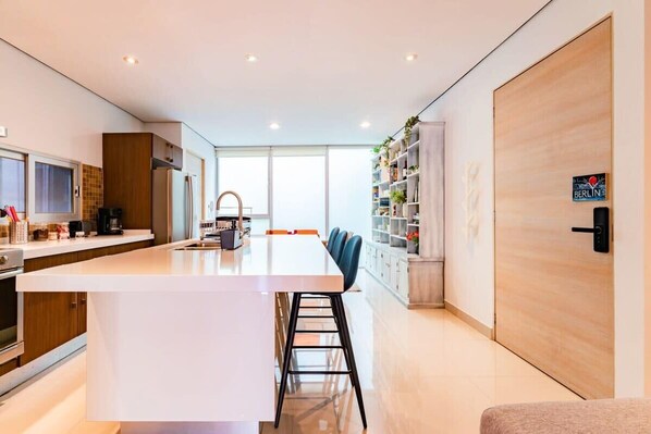 A modern kitchen with a central island, a dining area with chairs, and a bookshelf with plants.