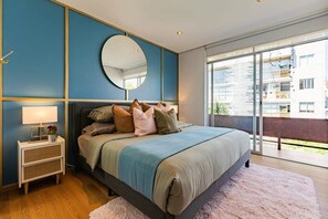 A bedroom with a large bed, a bedside table with a lamp, a round mirror, and a view of the outside.