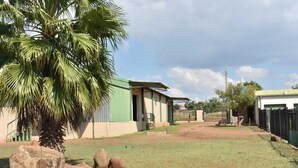 A building with a green roof and beige walls, surrounded by a grassy area with large rocks and a palm tree. - Sweetafrica lodge (Hartbeespoort)