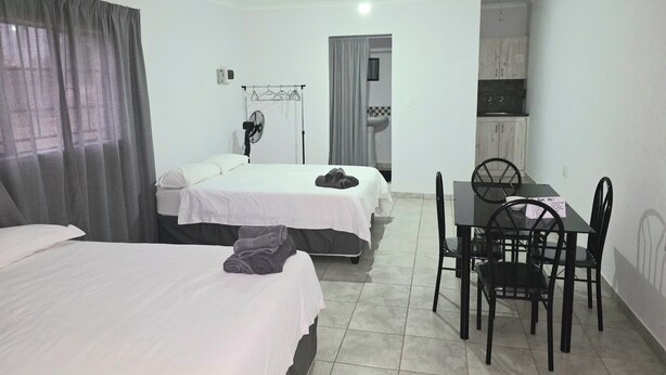 A hotel room with two beds, a dining table with chairs, a fan, and a kitchenette. - Sweetafrica lodge (Hartbeespoort)