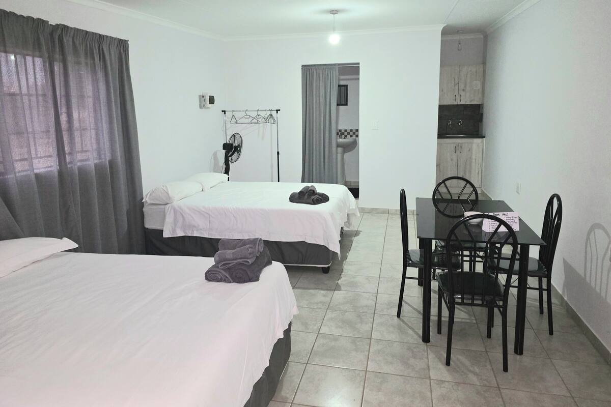 A hotel room with two beds, a dining table with chairs, a fan, and a kitchenette.