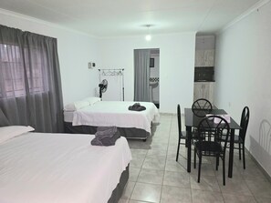 Een hotelkamer met twee bedden, een eettafel met stoelen, een ventilator en een kitchenette.