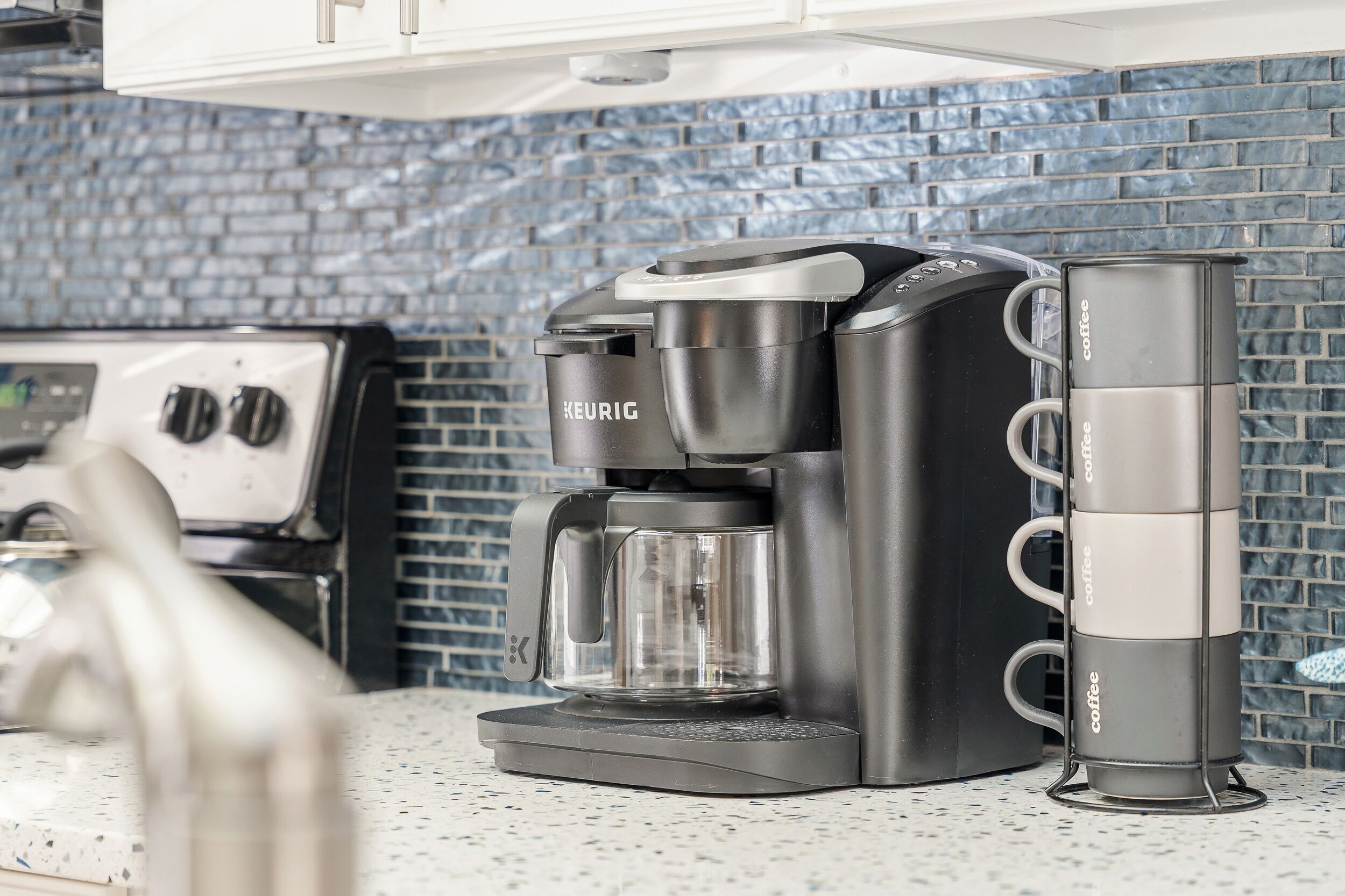 Une machine à café Keurig avec un porte-tasse à café à côté, posée sur un comptoir de cuisine dont le mur arrière est recouvert de carreaux.