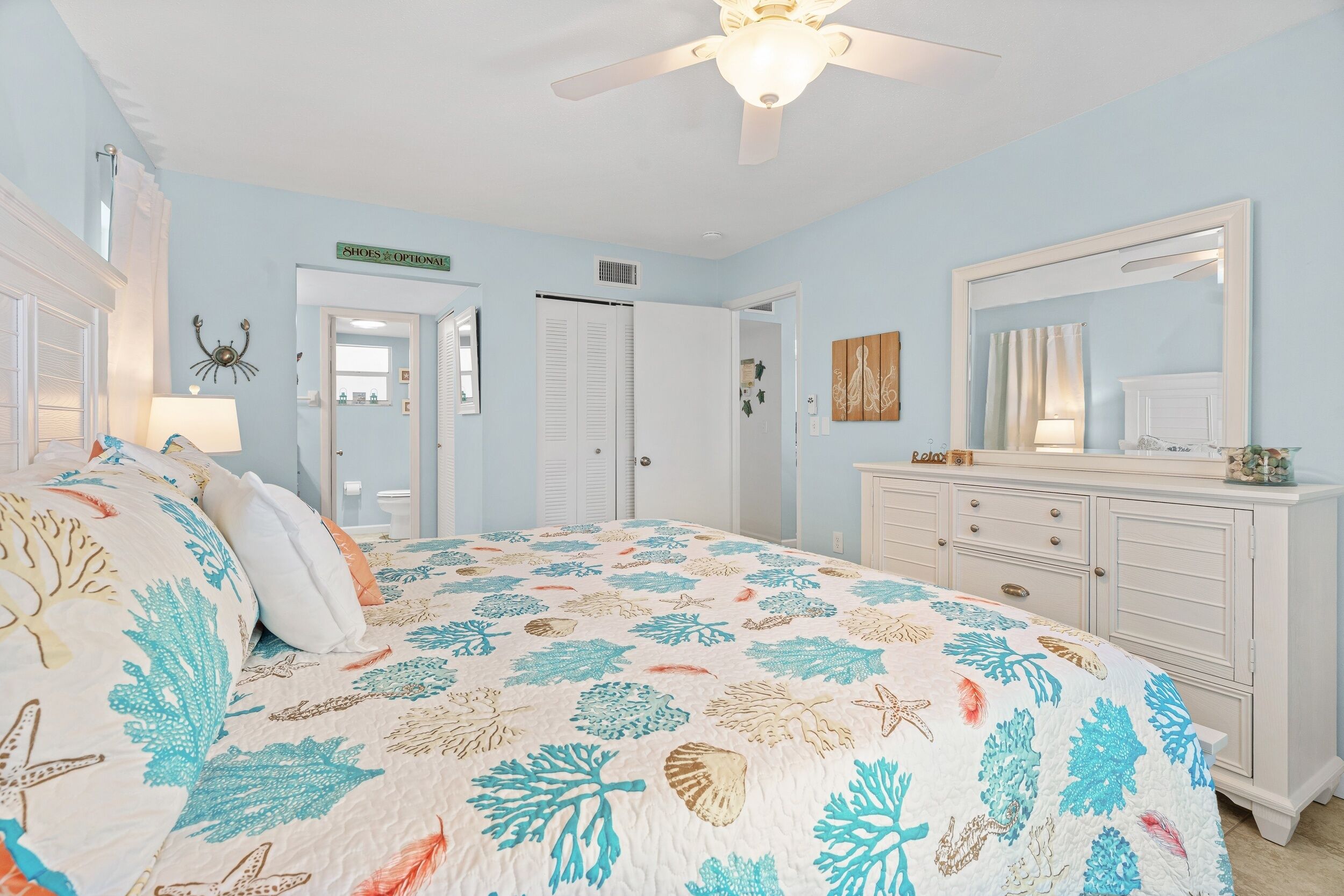 Une chambre à coucher comprenant un lit, une commode, un ventilateur de plafond et une porte donnant sur une autre pièce.