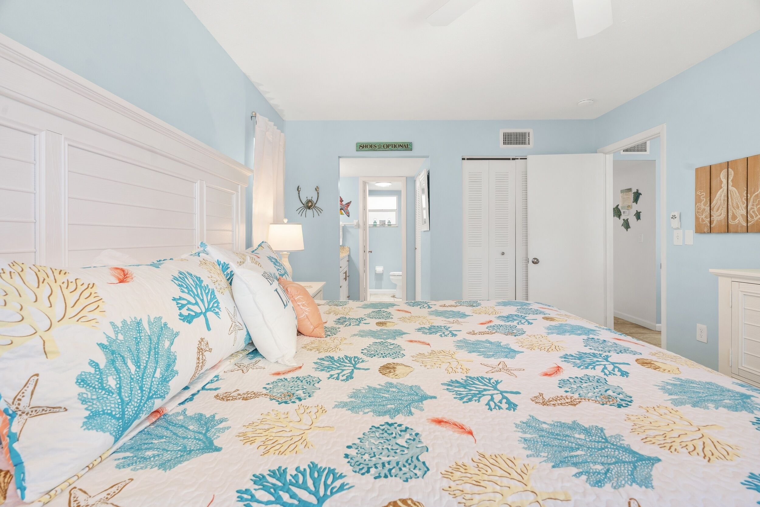 A bedroom with a bed covered in a coral-patterned quilt, a white headboard, and a door with a sign reading "SHOES OPTIONAL".