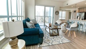 Una sala de estar moderna con sofá, una mesa de centro y sillas, una zona de cocina y ventanas amplias con vistas a la ciudad.