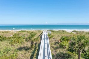 A beach with a wooden boardwalk leading to the ocean, surrounded by palm trees and coastal vegetation. - Oceanfront Complex | Heated Pool | Luxury Finishes (Cocoa Beach)