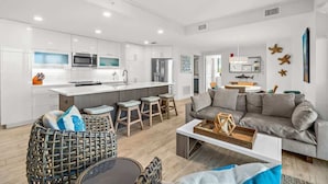 Una cocina moderna con armarios blancos, una isla central con taburetes, un comedor con un sofá gris y una sala de estar con un sofá gris y una silla de mimbre.