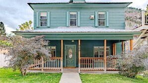A two-story house with a green exterior, a front porch with wooden railings, and a small deck on the right side. - Main Level Haven | Steps from the Hot springs | Sleeps 9 (Ouray)