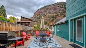 An outdoor patio with a grill, red chairs, and a table with an umbrella. A wooden fence and a mountain backdrop are visible. - Main Level Haven | Steps from the Hot springs | Sleeps 9 (Ouray)