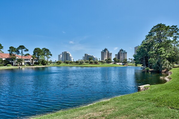 A lake with a grassy bank and trees, surrounded by buildings and a clear sky.