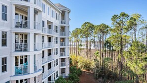 A multi-story hotel with balconies, a view of a forest, and a dirt path leading to the entrance.