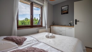 Un dormitori amb un llit gran, una finestra amb cortines, una tauleta de nit amb una planta i una porta.