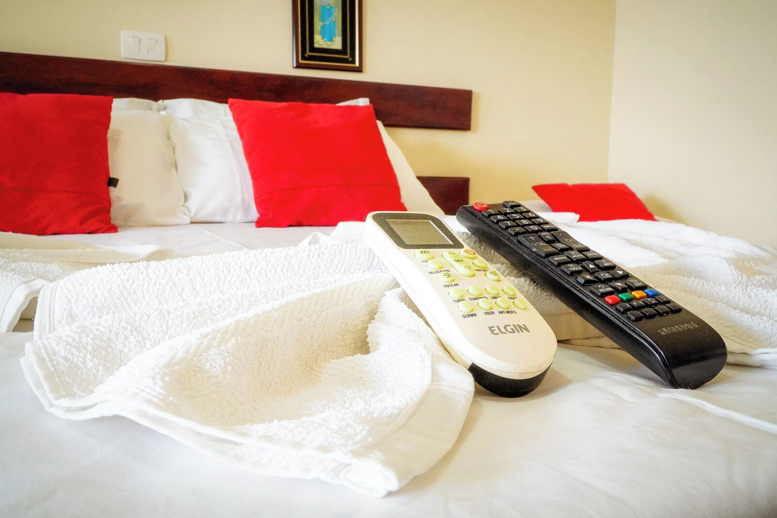 A bed with white and red pillows, a white Elgin remote control, and a black Samsung remote control on a white bedspread.