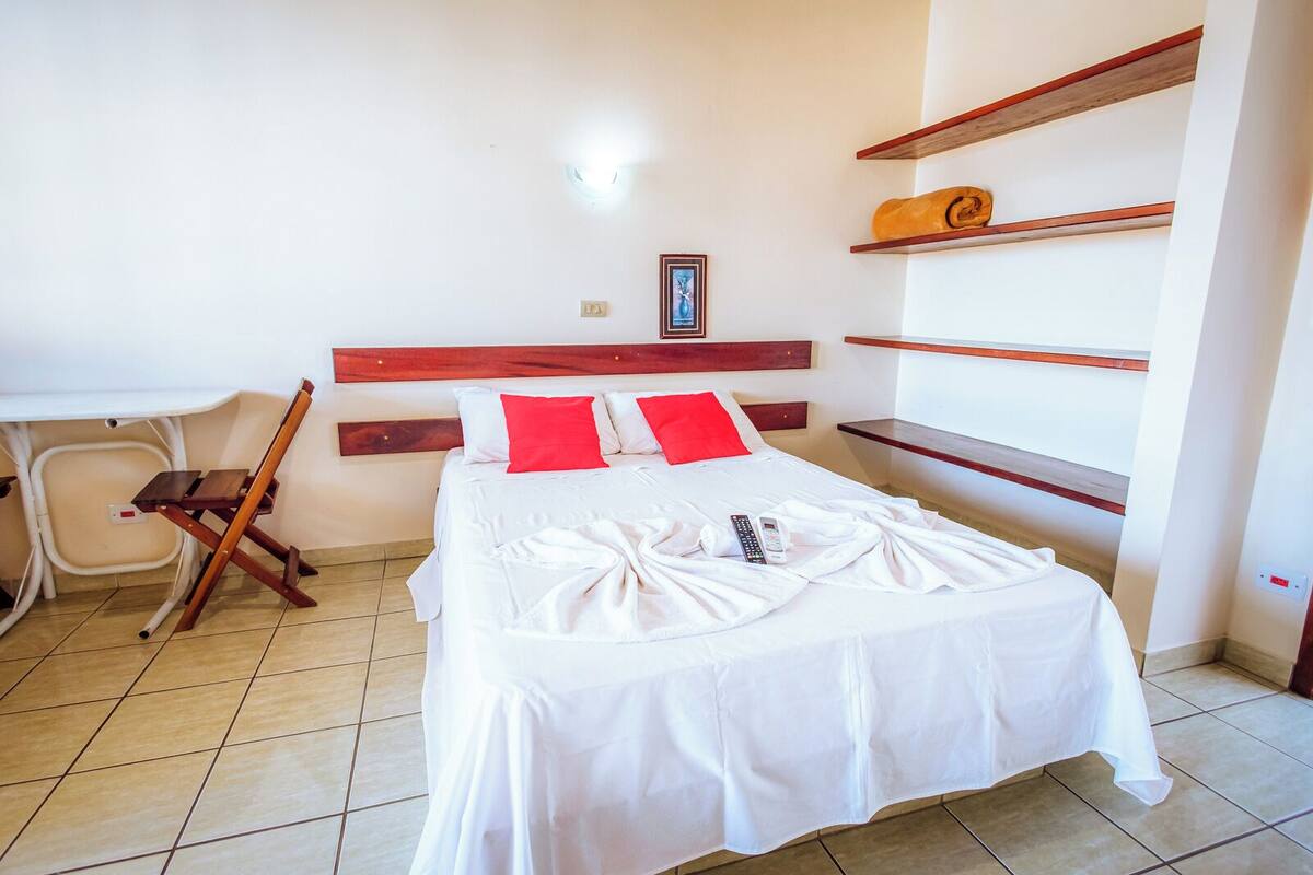 A neatly made bed with white sheets and red pillows, a wooden headboard, a small desk with a chair, and a shelf with a rolled-up towel.