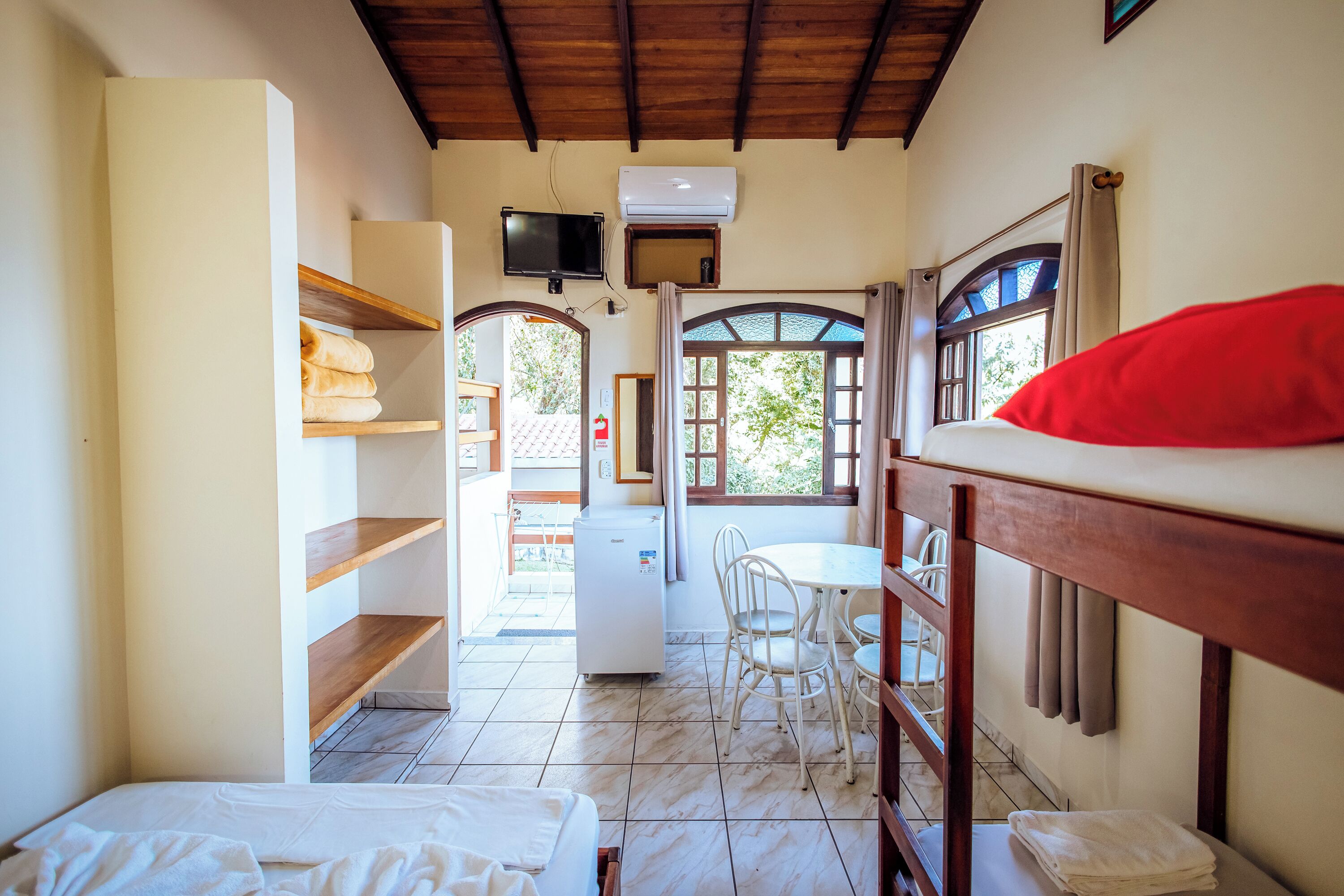A compact hotel room with a bunk bed, a small kitchenette, a dining area with a table and chairs, and a view through an arched window.