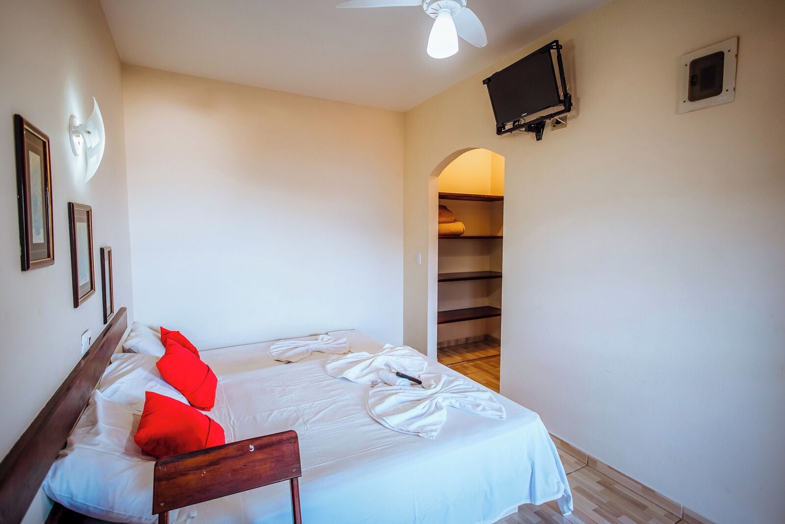 Une chambre à coucher comprenant un lit, une chaise en bois, un ventilateur de plafond, un téléviseur fixé au mur et une armoire avec des étagères.