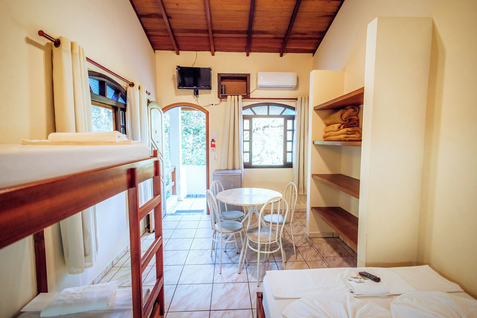 Quarto de hotel com teto de madeira, área de refeições com mesa e cadeiras, televisão fixada na parede e janela com cortinas.