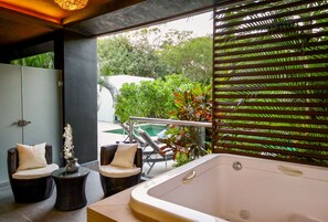 Un espace détente avec un jacuzzi, deux fauteuils en osier dotés de coussins, une petite table ornée d’un vase de fleurs, et une vue sur une piscine et la verdure environnante.