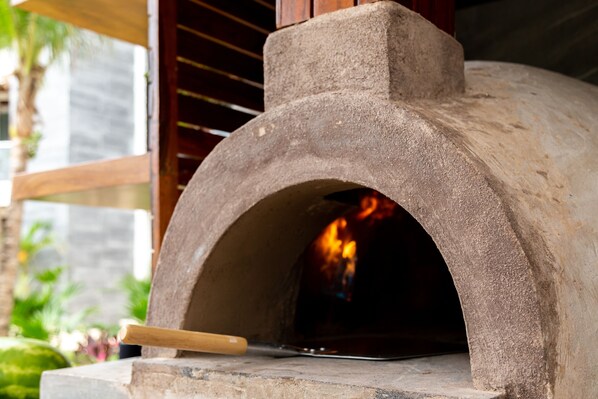 Un four à pizza extérieur en pierre, doté d’une cheminée, d’une pelle à pizza et d’une structure en bois en arrière-plan.
