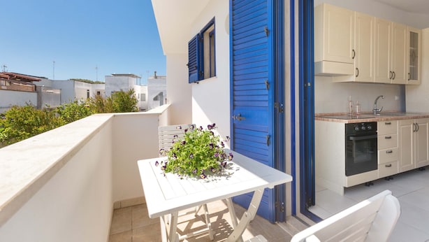 A balcony with a table and chairs, a kitchen with blue shutters, and a view of neighboring buildings. - 3403 Casa Petrachi - PP by Barbarhouse (Melendugno)