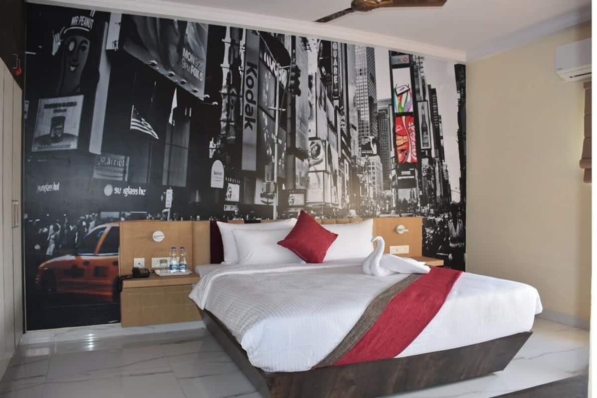 A hotel room with a large bed, a wooden headboard, a nightstand with a lamp, and a wall mural of a cityscape.