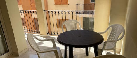 Un balcón con una mesa redonda y cuatro sillas, barandilla y vista a un edificio con persianas de madera.