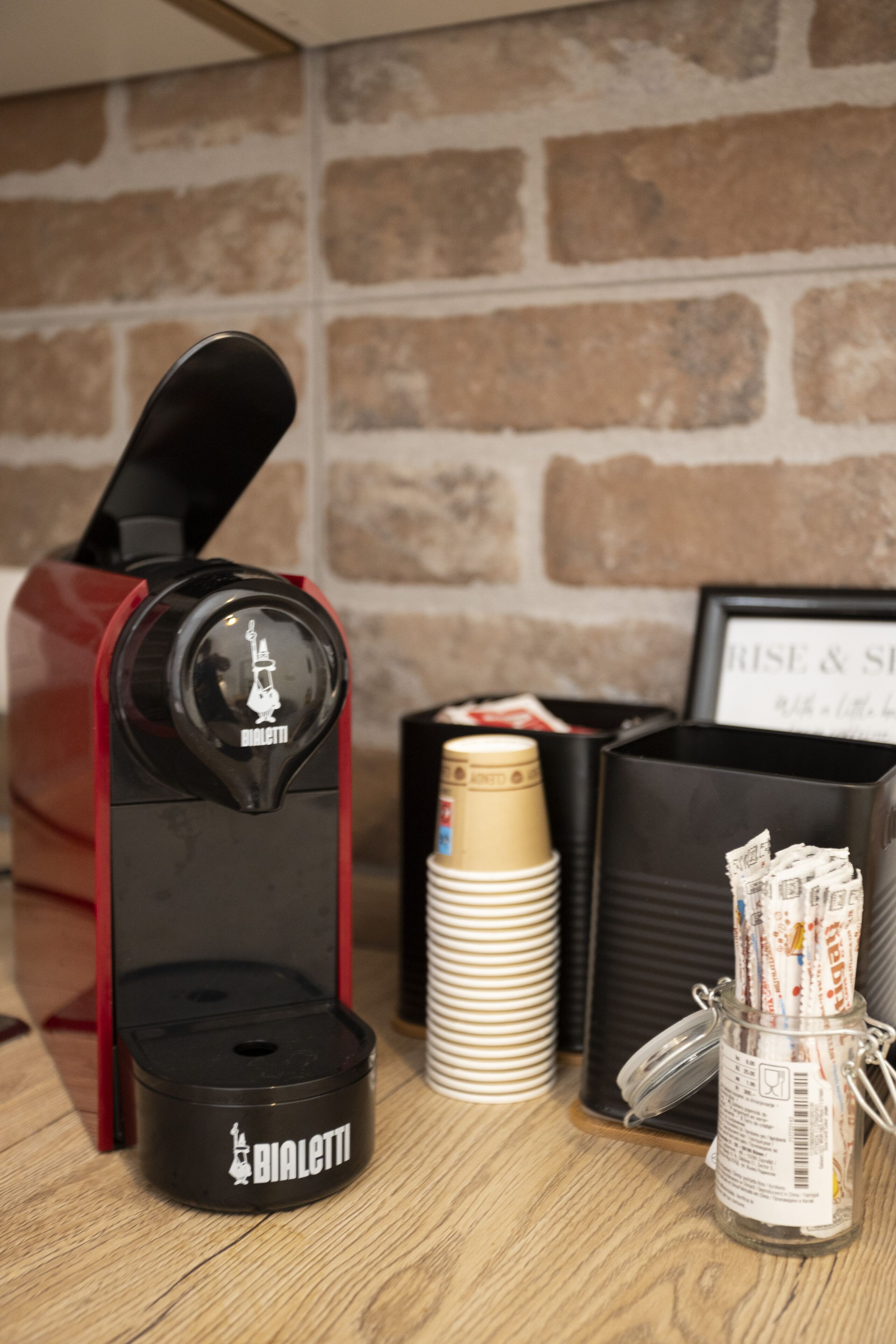 Une machine à café, une pile de gobelets jetables et un bocal en verre contenant des sachets, probablement du sucre ou de la crème liquide, sont posés sur une table en bois, adossée à un mur de briques.