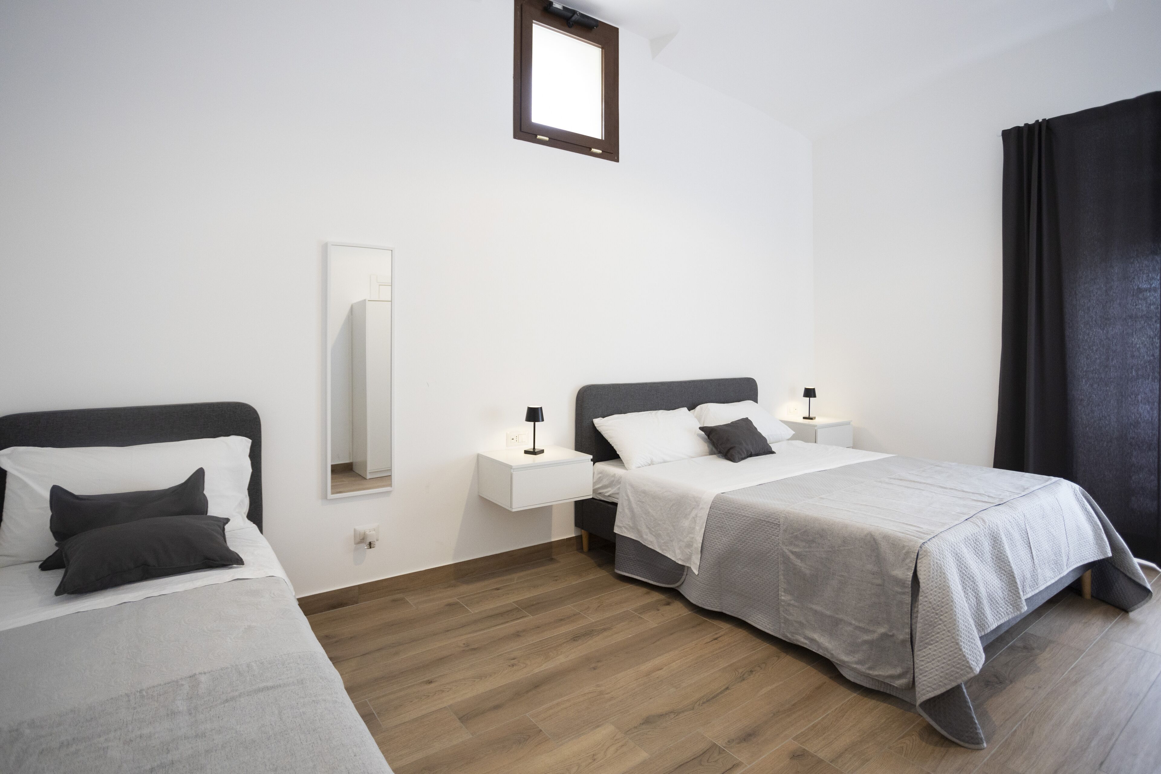 Una habitación de hotel moderna con dos camas individuales, una mesita de noche pequeña con lámpara, un espejo grande y una ventana con cortinas.