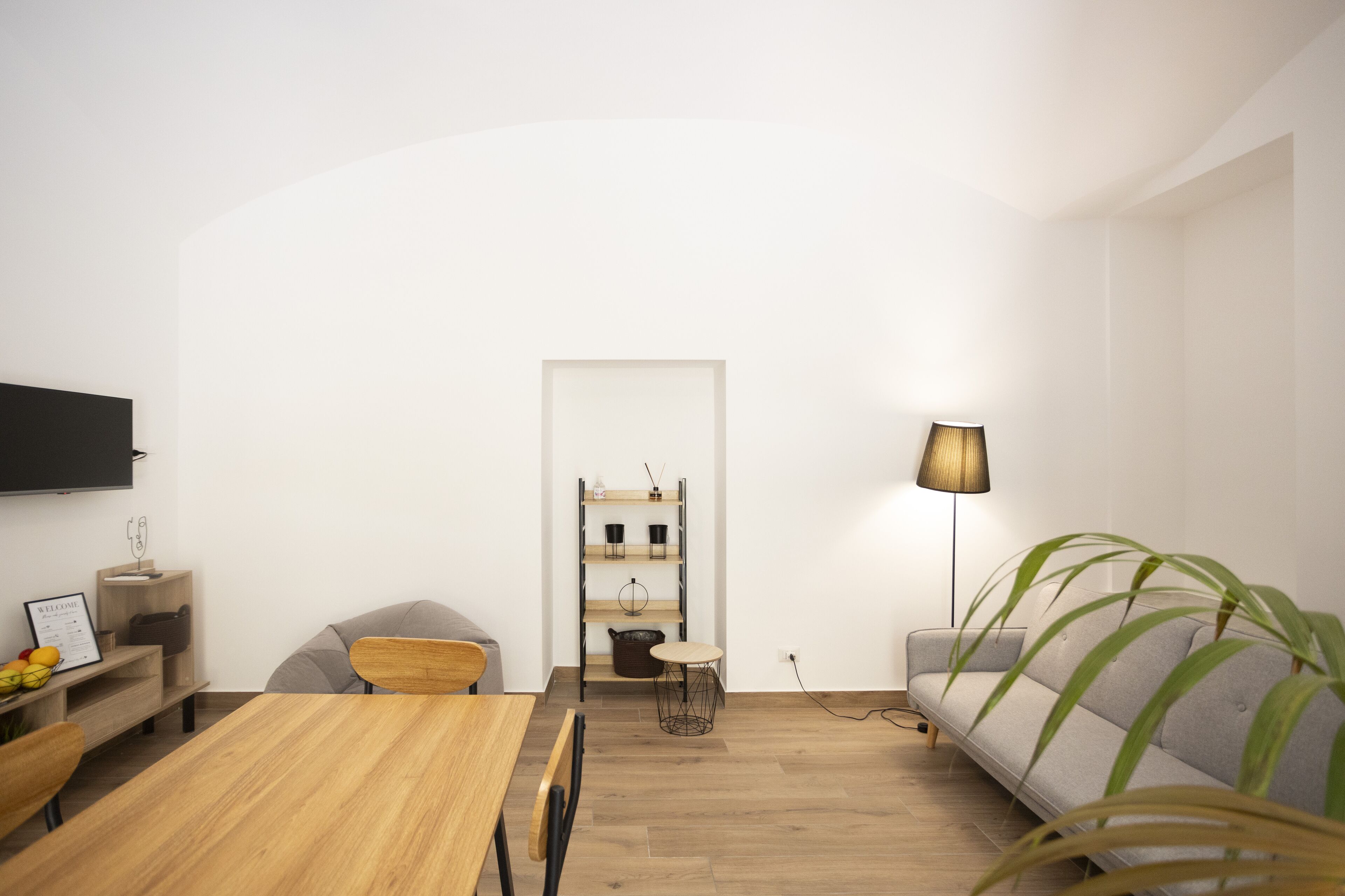 A modern, minimalistic living room with a wooden dining table, grey sofa, and a TV mounted on the wall.