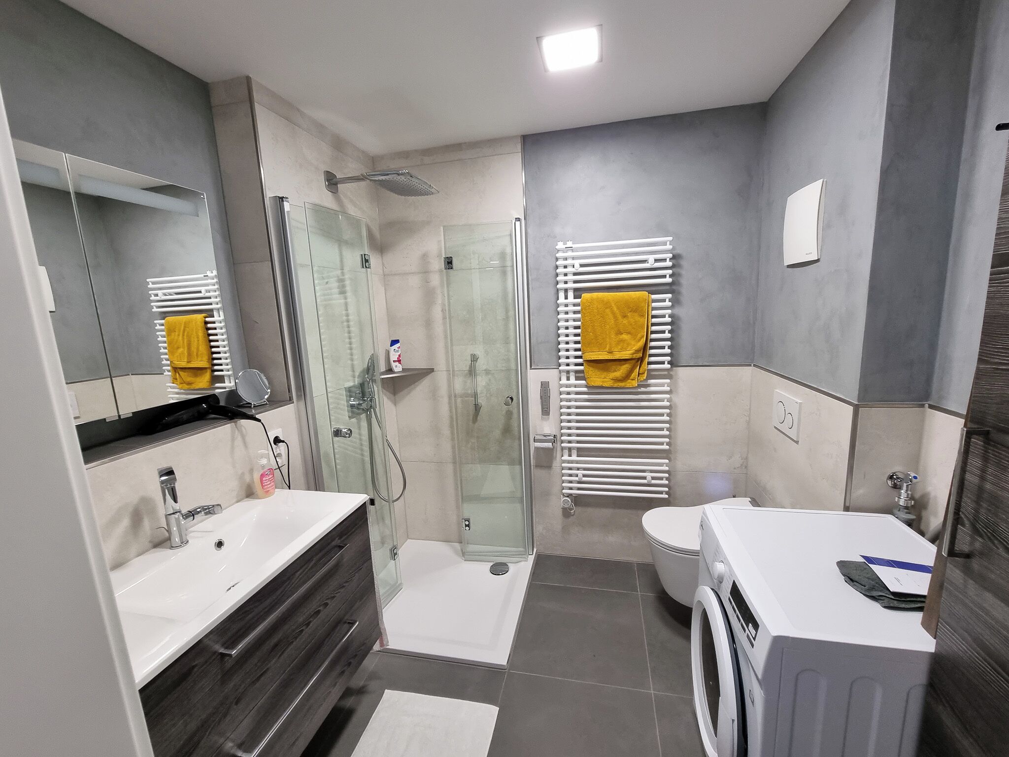 A modern bathroom with a glass-enclosed shower, a white sink, a toilet, and a built-in washing machine.