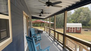 A covered porch with blue rocking chairs, a ceiling fan, and a view of a red-roofed building in the background. - 2 bed, 2 bath cabin located in the country just west of College Station.   (Caldwell)