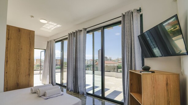 A hotel room with a large bed, a wooden sideboard, a flat-screen TV, and a view of the city through floor-to-ceiling windows. - Vullkan Hotel (Ksamil)