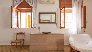 Ein Zimmer mit einem Holzkoffer, einem Stuhl, einem Bett und zwei Fenstern mit Vorhängen.