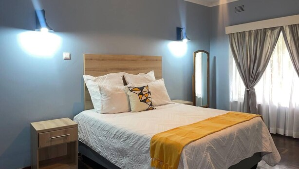 Een slaapkamer met een houten hoofdbord, een bed met witte beddengoed en een gele plaid, twee bedlampen, een nachtkastje en een raam met gordijnen.