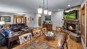 A spacious living room with a fireplace, leather sofas, a dining table with chairs, and a large TV mounted on the wall. - 4-Bedroom Mountain House w/Golf Course View, Hot Tub, Pool Table, & Foosball! (Whittier)