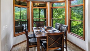 A dining area with a table set for six, surrounded by large windows offering a view of a forested landscape. - 4-Bedroom Mountain House w/Golf Course View, Hot Tub, Pool Table, & Foosball! (Whittier)